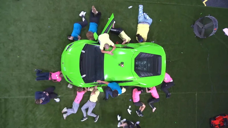 Overhead view of a video set: contestants in bright shirts lying around a green sports car on artificial turf